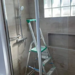 Montaż kabiny prysznicowej z szybą i szklanymi luksferami w oknie, drabina z zielonymi elementami wewnątrz kabiny, widoczne ślady fugowania na płytkach i drabince.