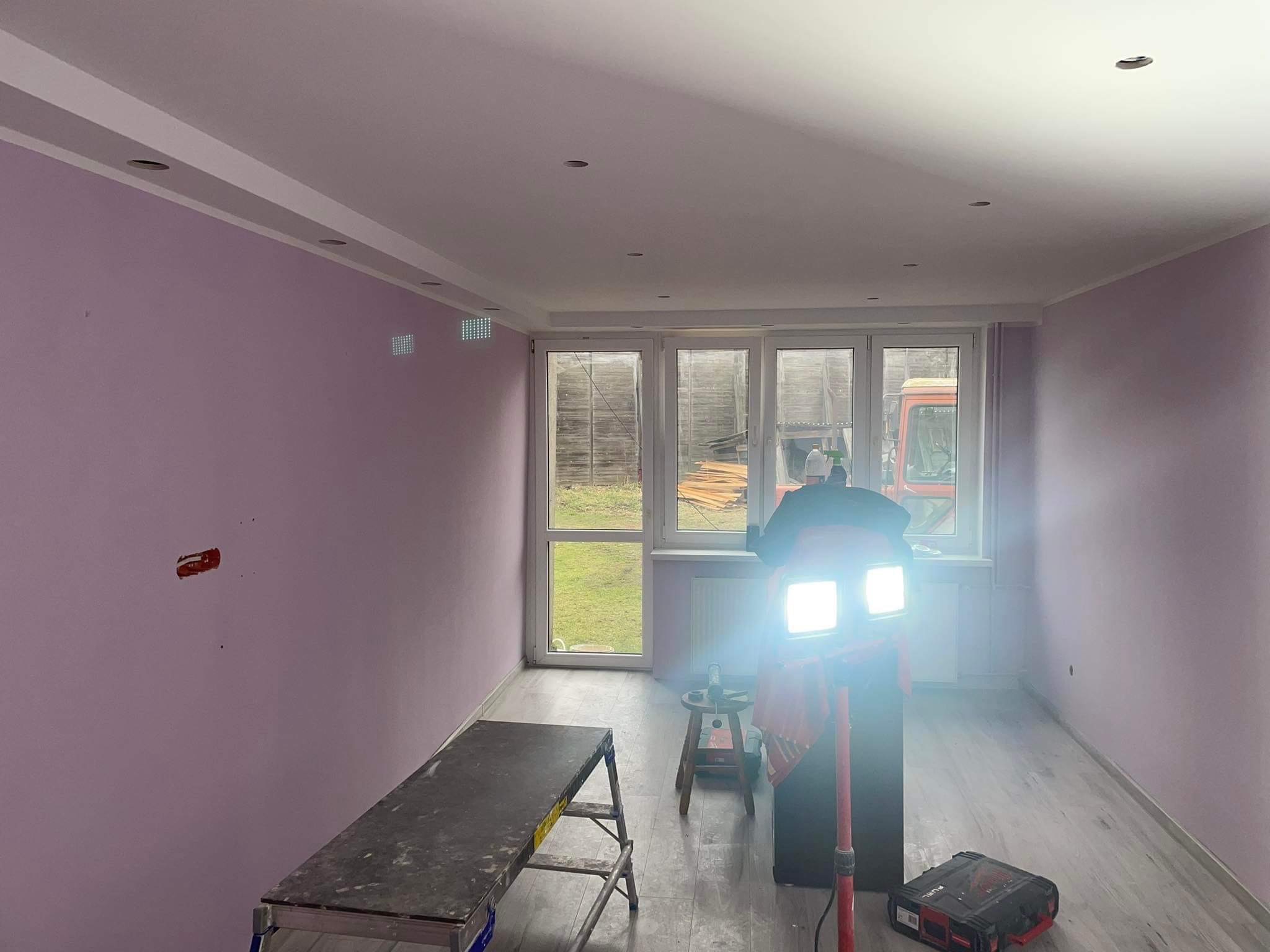 Pokój po malowaniu na delikatny fiolet, z widoczną instalacją elektryczną, drabina, lampa budowlana i skrzynka narzędziowa, okno z widokiem na zewnątrz.