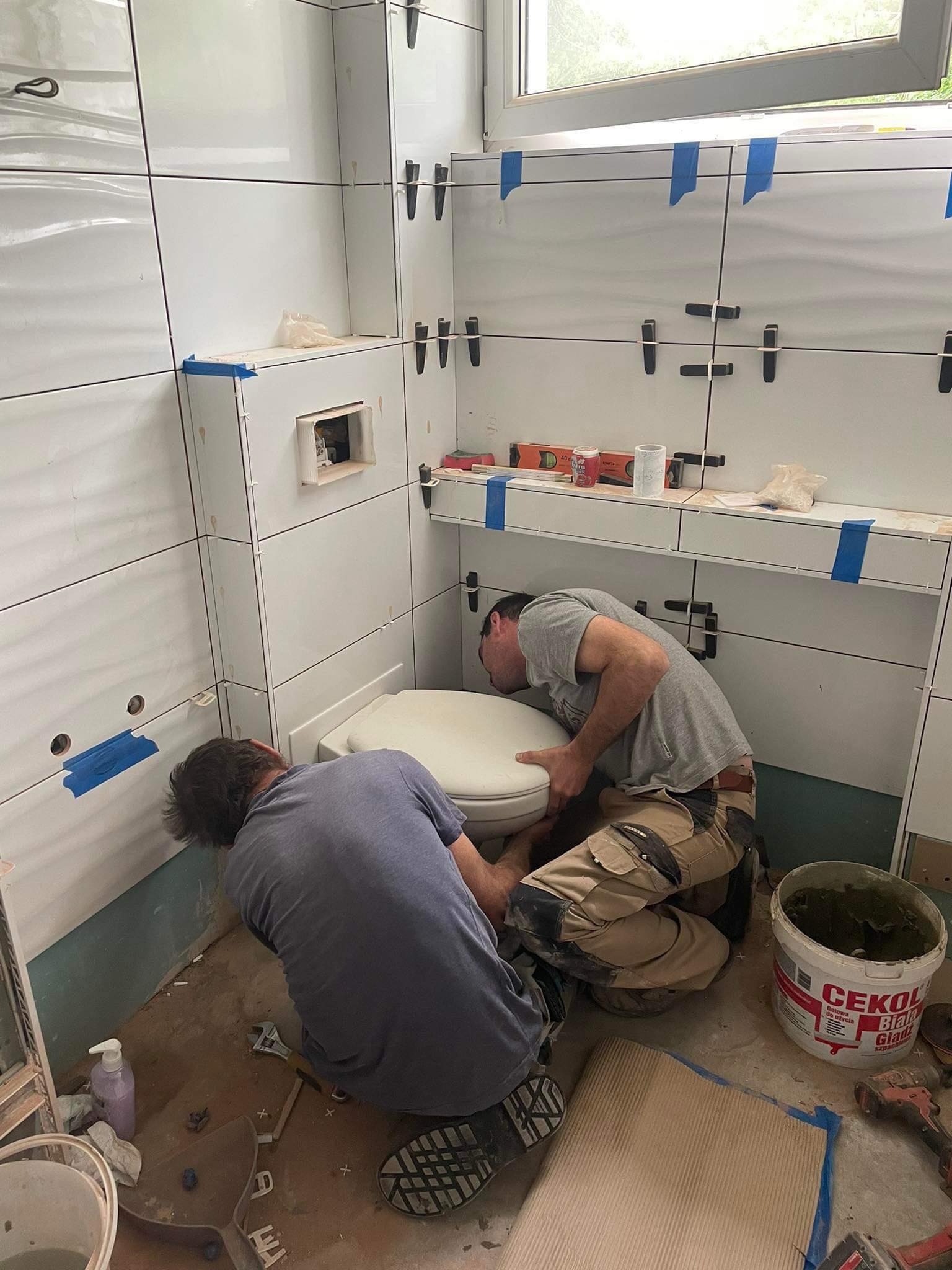 Dwóch mężczyzn instaluje kompakt WC w łazience z białymi, błyszczącymi płytkami, widoczne narzędzia i wiadro z zaprawą Cekol.