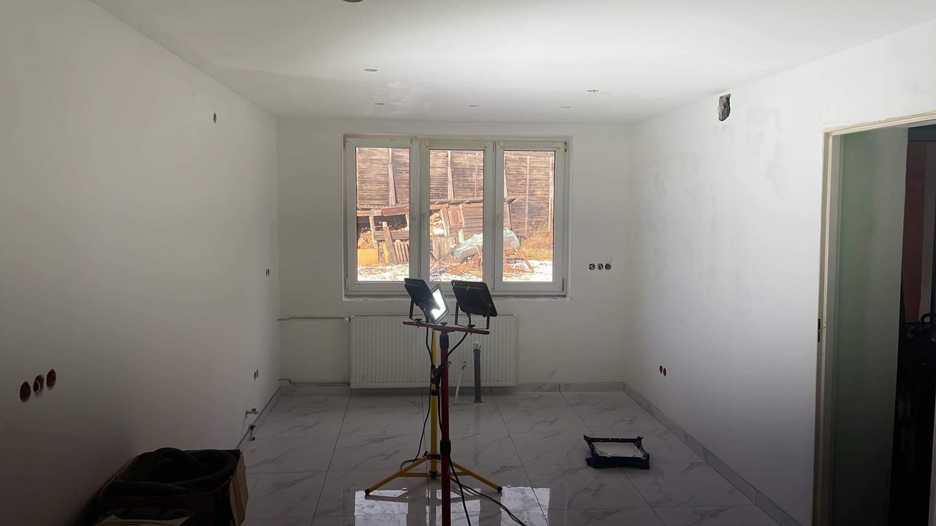 Pokój po malowaniu ścian na biało, widoczne gniazdka elektryczne, marmurowe płytki na podłodze i lampy budowlane oświetlające wnętrze, okno z widokiem na zewnątrz.