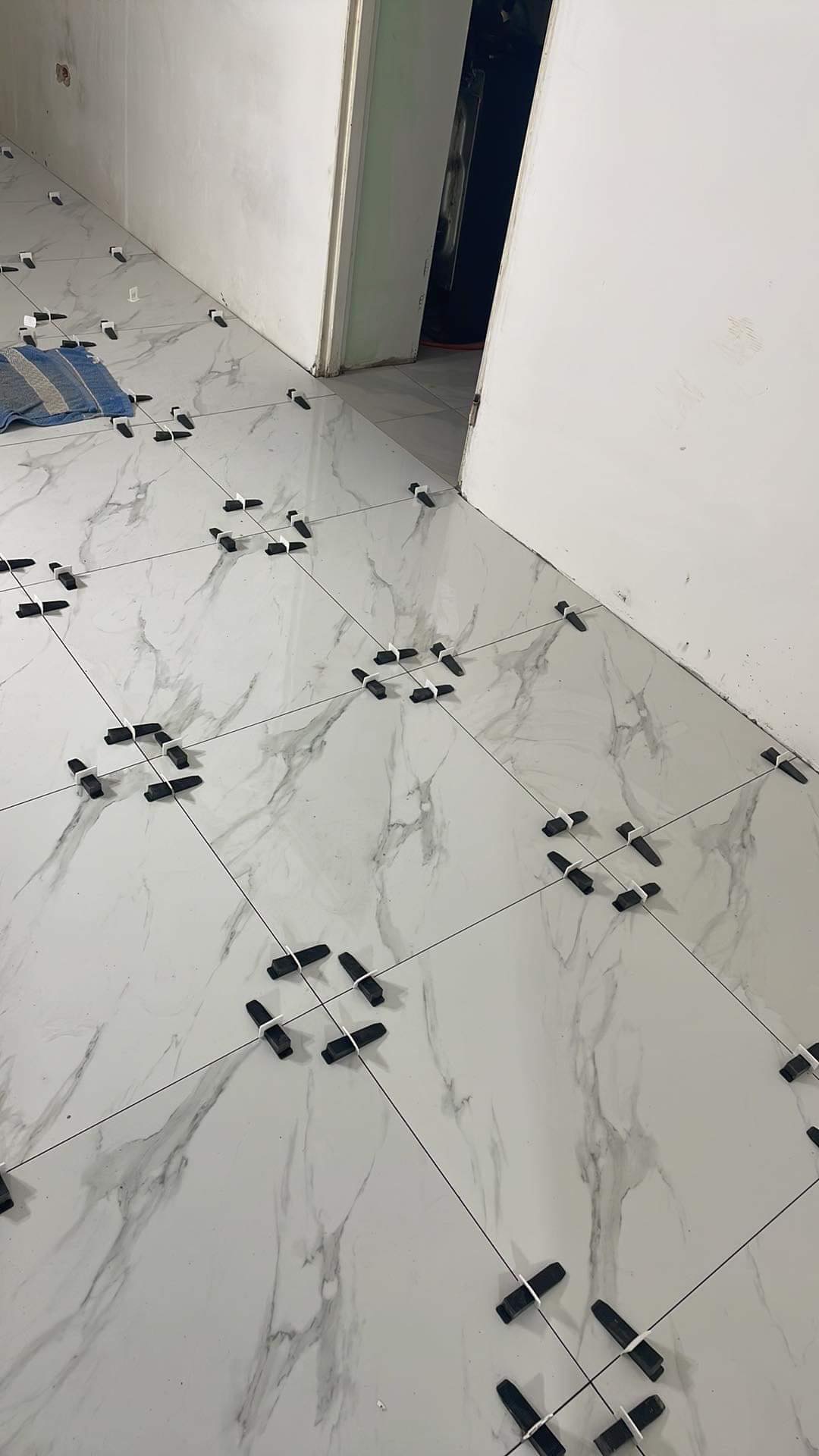 Układanie białych płytek imitujących marmur z użyciem czarnych krzyżyków dystansowych i poziomowania podłogi w trakcie remontu.