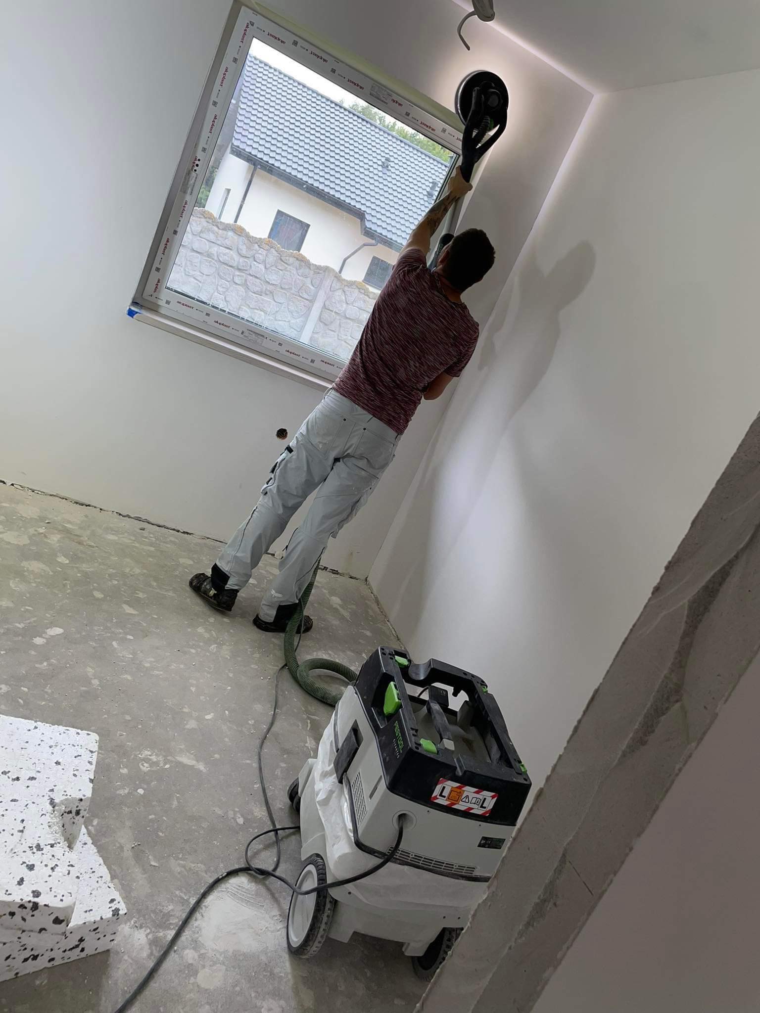 Pracownik szlifuje ścianę w pomieszczeniu z oknem, używając szlifierki z odkurzaczem przemysłowym Festool, widoczne ślady szlifowania na ścianie i pył na podłodze.