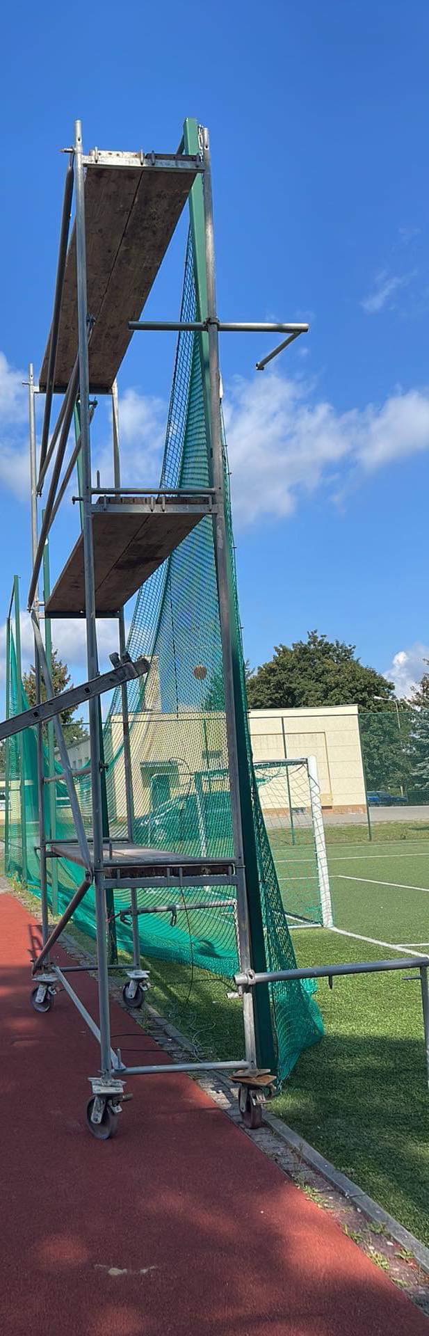 Rozstawione rusztowanie na kółkach z siatką ochronną, ustawione na czerwonej bieżni sportowej, w tle zielone boisko i budynek.