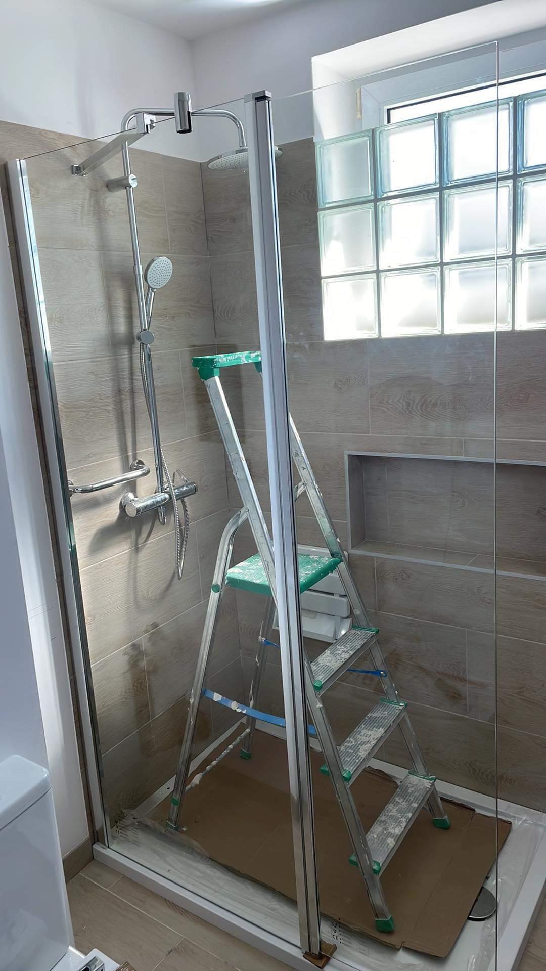 Montaż kabiny prysznicowej z szybą i szklanymi luksferami w oknie, drabina z zielonymi elementami wewnątrz kabiny, widoczne ślady fugowania na płytkach i drabince.