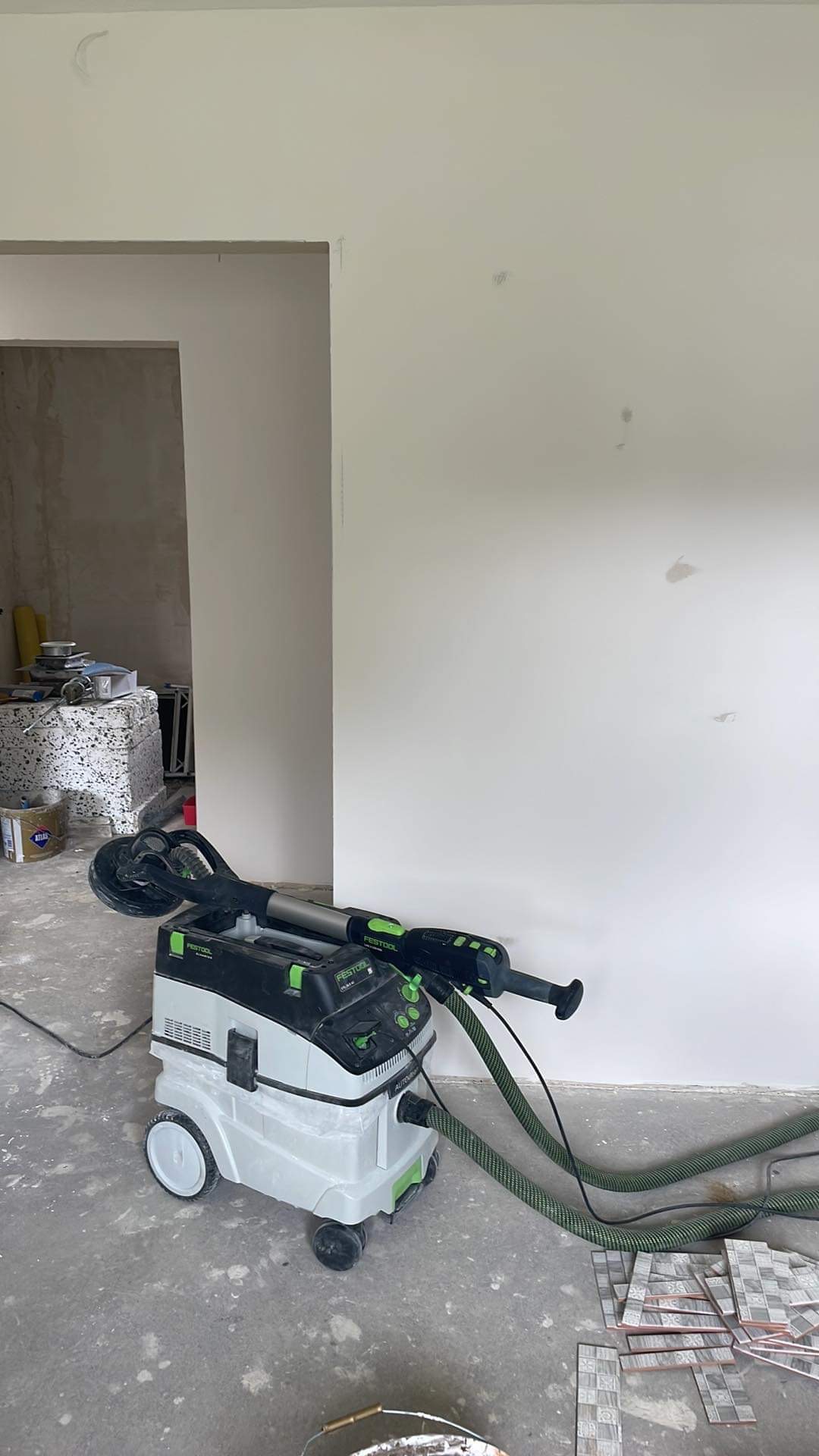 Szlifierka Festool Planex LHS 225 EQ z odkurzaczem stojąca w pomieszczeniu w trakcie prac wykończeniowych, widoczne ślady szpachli na ścianie i podłodze, w tle materiały budowlane.