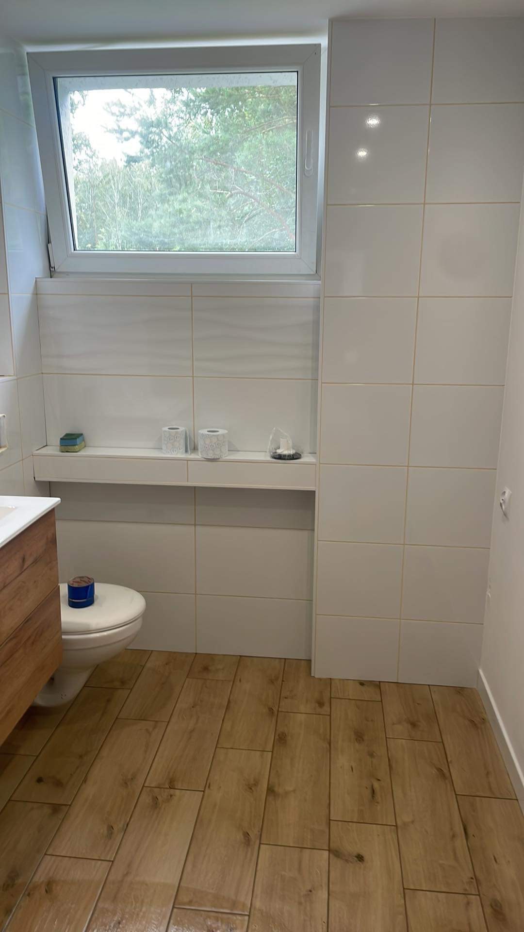 Białe płytki ścienne i drewnopodobne płytki podłogowe w łazience z oknem, półka w zabudowie z akcesoriami toaletowymi, fragment szafki pod umywalką z drewnianym frontem.