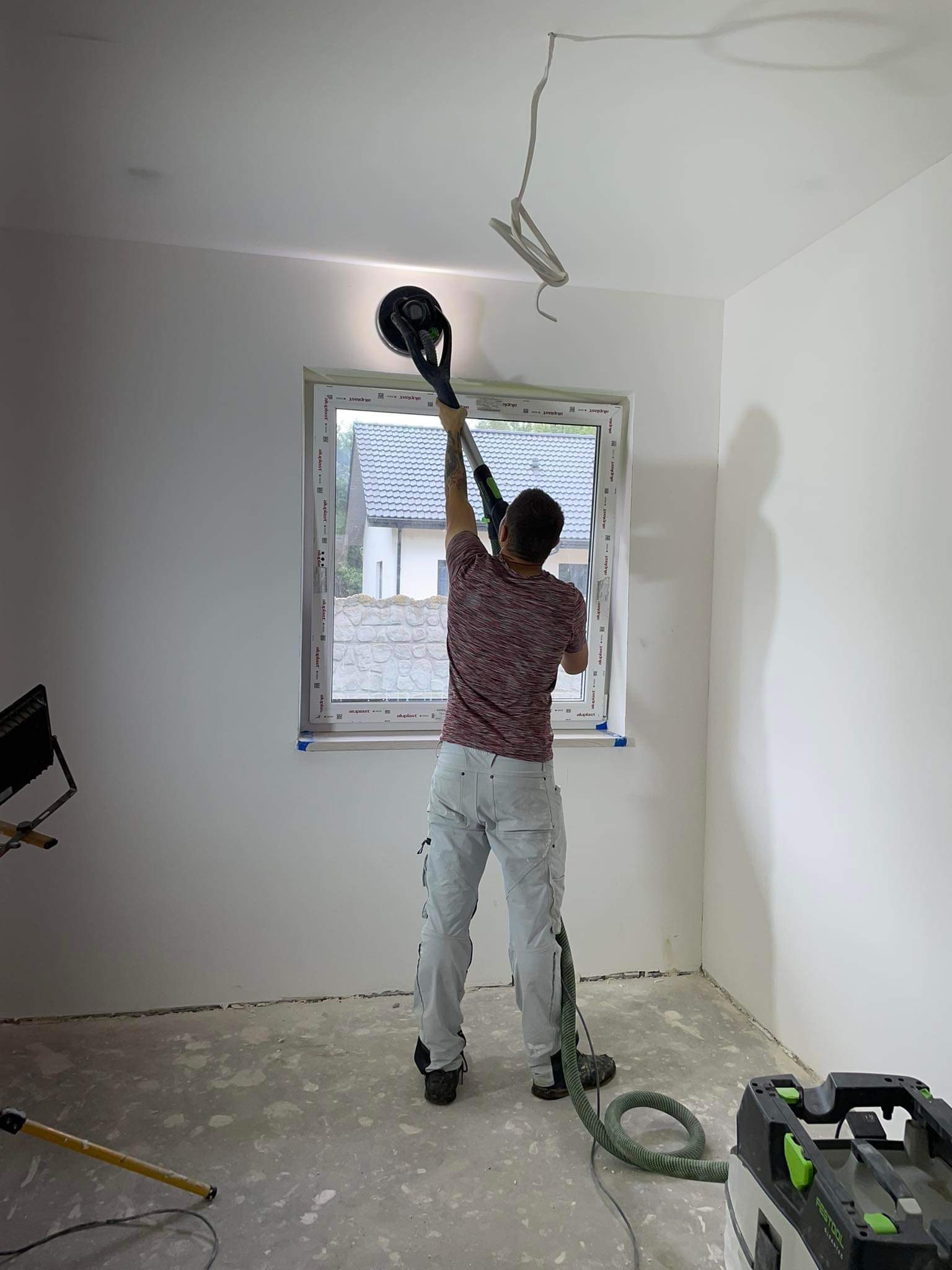 Pracownik szlifuje sufit gładzią szpachlową za pomocą szlifierki z odkurzaczem Festool, widoczne okno z widokiem na dom i wiszące kable.