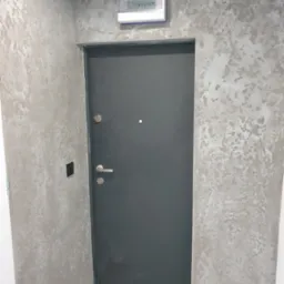 Ściana pokryta dekoracyjną szarą gładzią o nieregularnej fakturze, widoczna szara, nowoczesna stolarka drzwiowa z metalową ościeżnicą oraz biała skrzynka elektryczna nad drzwiami.