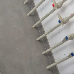 Szare płytki podłogowe ułożone w rzędzie, obok instalacji wodnej z rurami i zaworami z kolorowymi oznaczeniami (czerwonymi i niebieskimi)