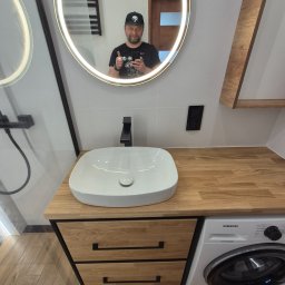 My House EWELINA WINIARSKA - Nowoczesna łazienka z umywalką nablatową, drewnianym blatem i szafką z szufladami. W tle lustro z podświetleniem LED i odbicie osoby.