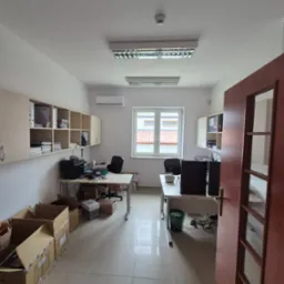 Jasne biuro po remoncie: białe ściany, płytki podłogowe, biurka z komputerami, kartony do przeprowadzki, klimatyzacja, otwarte drzwi. Widok z perspektywy wejścia.