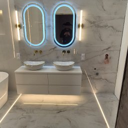 My House EWELINA WINIARSKA - Nowoczesna łazienka z dwoma owalnymi lustrami LED, białymi umywalkami nablatowymi i marmurowymi płytkami. Widoczne złote detale armatury i oświetlenia.