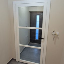 My House EWELINA WINIARSKA - Białe, przeszklone drzwi wewnętrzne z trzema poziomymi szprosami, widoczne na tle szarej podłogi i jasnych ścian. W głębi widoczny fragment kolejnych drzwi.