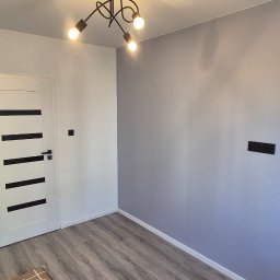 Pokój po remoncie: szare ściany, drewnopodobna podłoga, białe drzwi z czarnymi paskami i nowoczesna lampa sufitowa. Widoczne gniazdka elektryczne.