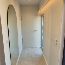 My House EWELINA WINIARSKA - Jasny korytarz z beżowymi płytkami podłogowymi i minimalistycznym lustrem w kształcie łuku. Sufit z oświetleniem punktowym i ukrytym paskiem LED.