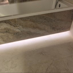 My House EWELINA WINIARSKA - Wykończona łazienka z wanną obudowaną płytkami imitującymi marmur i podświetleniem LED. Widoczne przyłącza wodne na ścianie. Ukośne ujęcie.