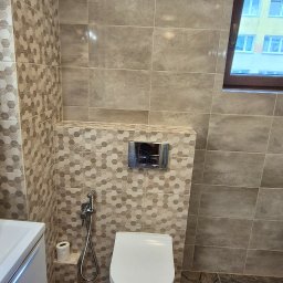 My House - Wykończona łazienka z podwieszaną toaletą, mozaiką heksagonalną i płytkami imitującymi marmur na podłodze. Widok na okno z budynkiem w tle.
