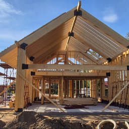 My House - Konstrukcja domu w trakcie budowy, drewniany szkielet z widocznymi elementami łączeń, rusztowania i materiały budowlane na placu budowy w słoneczny dzień.