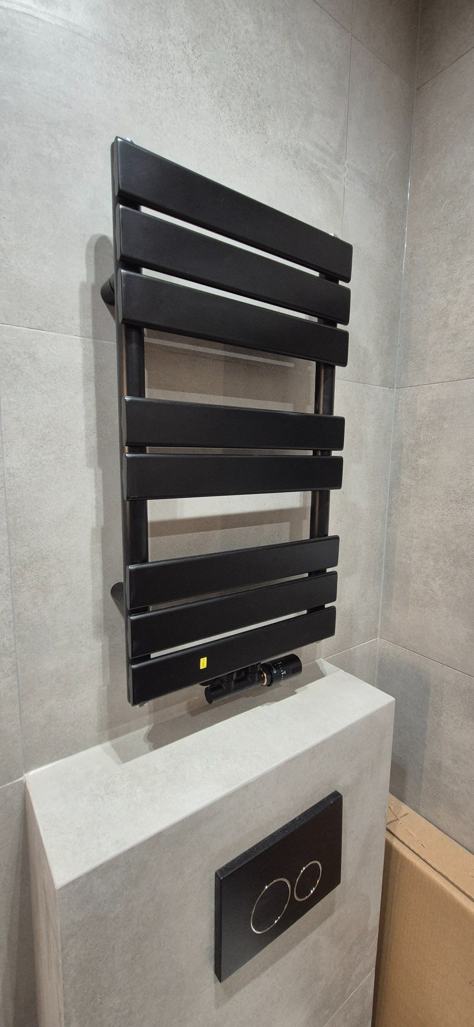 Czarny, drabinkowy grzejnik łazienkowy na tle szarych płytek. Nowoczesny design, minimalistyczny styl. Widoczny fragment zabudowy WC z czarnym przyciskiem.