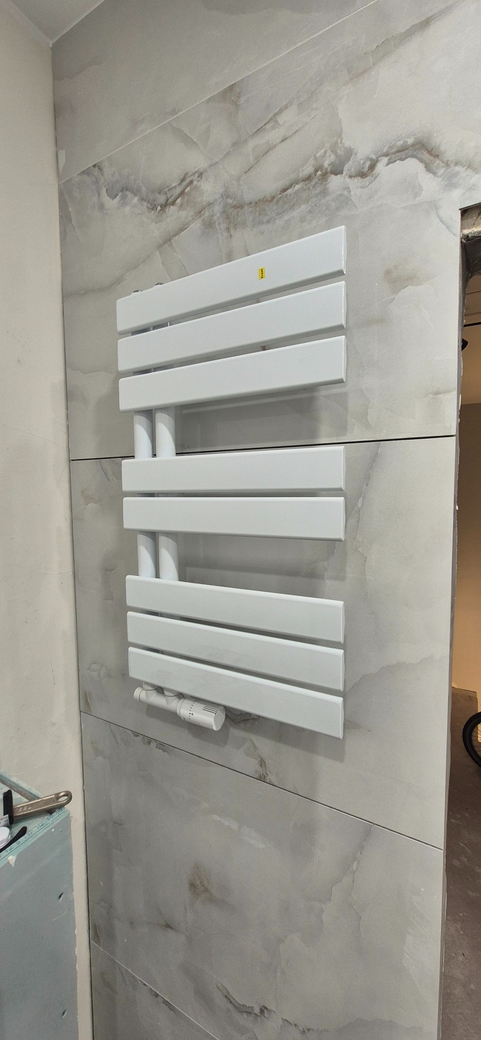 Biały grzejnik drabinkowy zamontowany na ścianie wyłożonej płytkami imitującymi marmur. Widoczny fragment narzędzi i płyty gipsowej, sugerujący etap wykończenia łazienki.