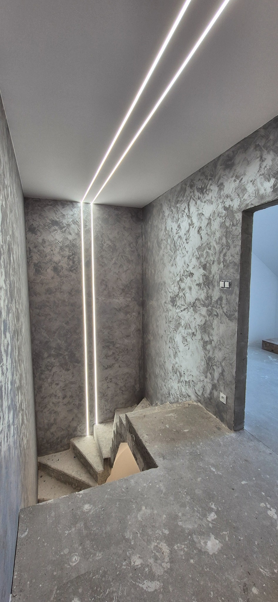 Klatka schodowa w szarościach z dekoracyjną strukturą ścian i nowoczesnym oświetleniem LED. Widoczne surowe schody i fragment otwartego przejścia.