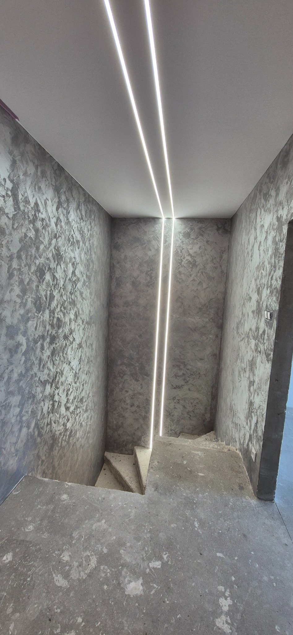 Klatka schodowa w szarościach z dekoracyjną strukturą na ścianach i nowoczesnym oświetleniem LED w suficie. Widok z dołu, surowy stan wykończenia podłogi.