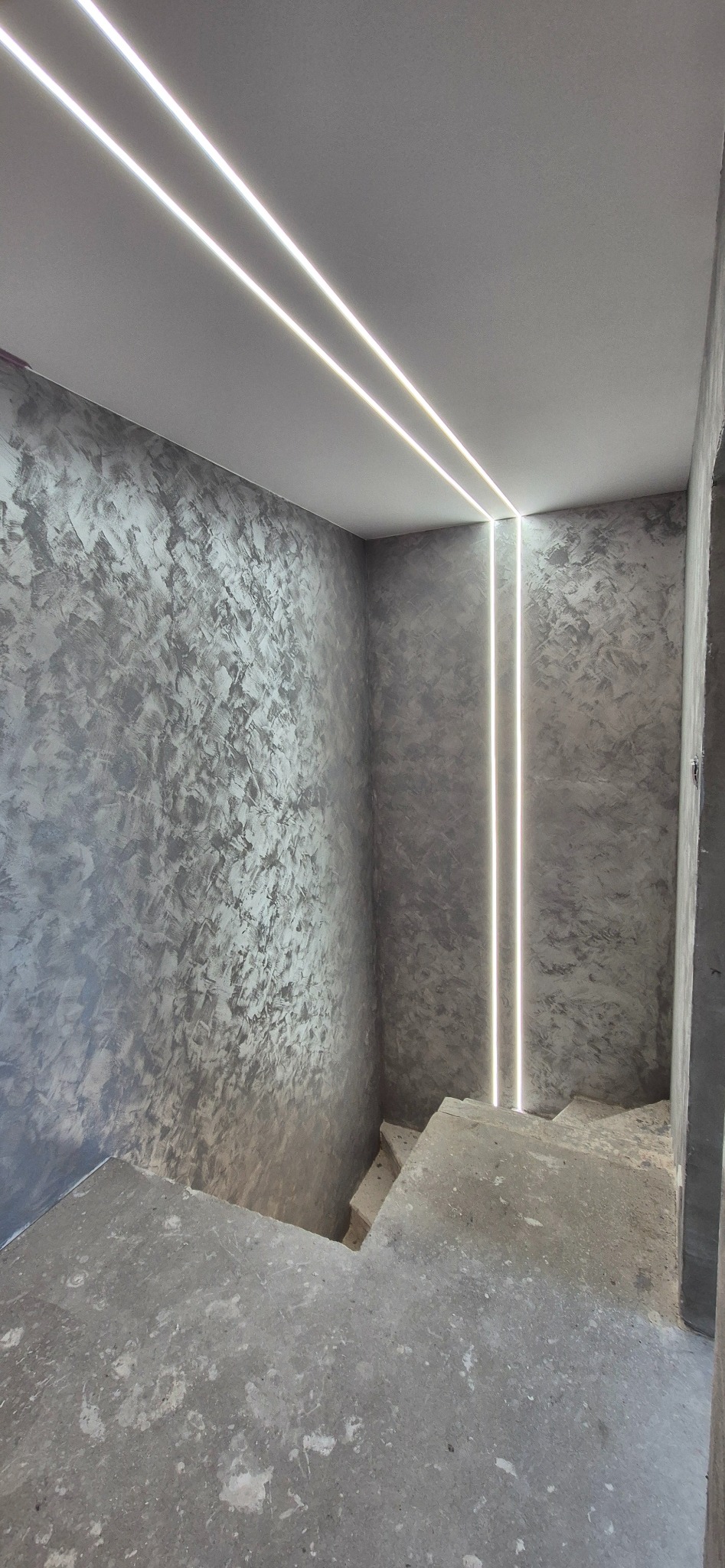 Szara ściana ze strukturą i oświetleniem LED na suficie, klatka schodowa w surowym stanie. Minimalistyczny design wnętrza w trakcie wykańczania.