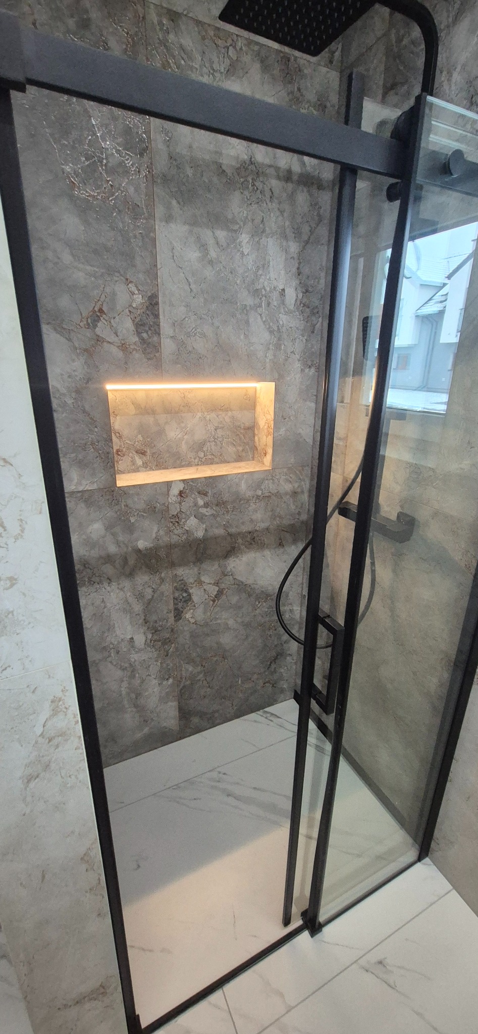 Nowoczesna kabina prysznicowa z czarnymi profilami i marmurowymi płytkami. W ścianie wnęka z podświetleniem. Biała marmurowa podłoga. Minimalistyczny design.