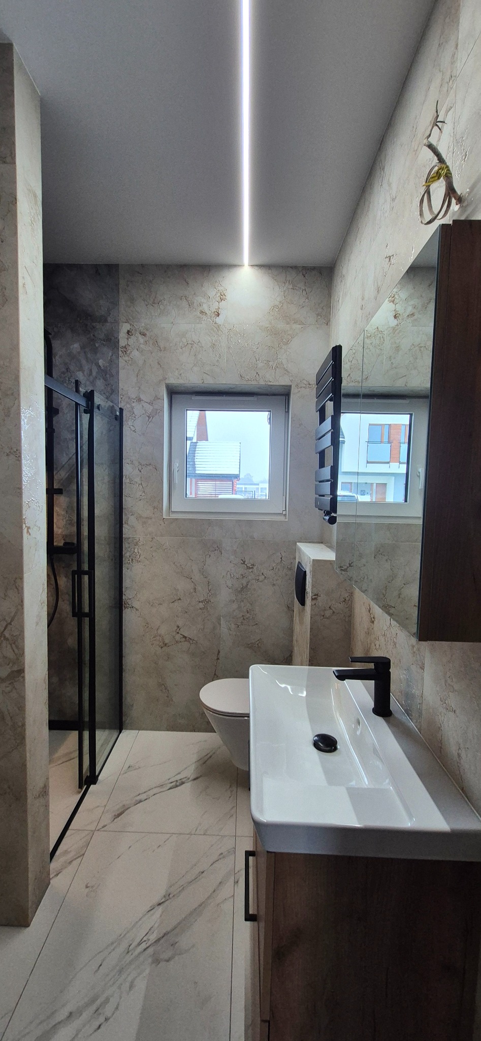 Nowoczesna łazienka po remoncie: kabina prysznicowa z czarnymi ramami, umywalka z szafką w kolorze drewna, marmurowe płytki i podłużne oświetlenie LED.