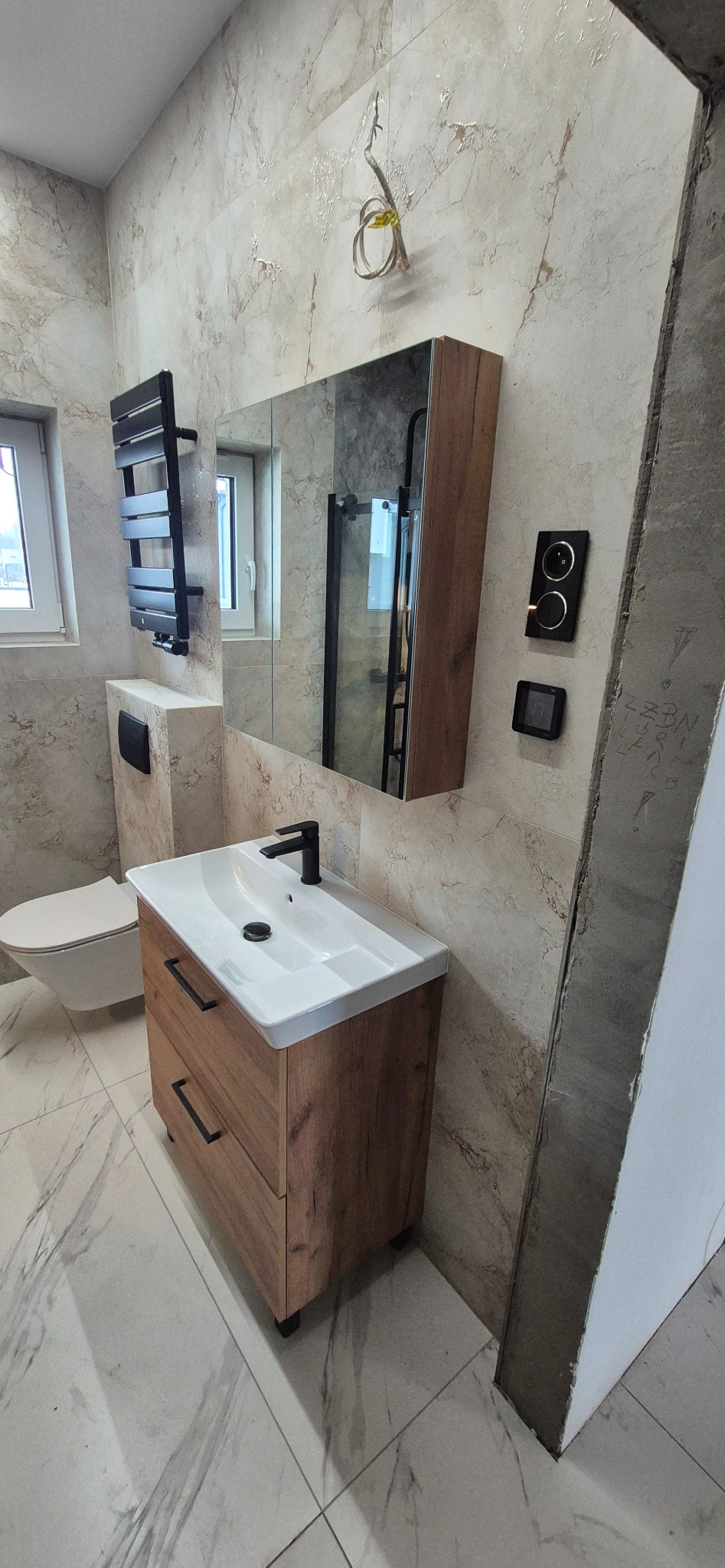 Nowoczesna łazienka po remoncie: drewniana szafka z umywalką, lustro z oświetleniem, grzejnik drabinkowy i marmurowe płytki. Widoczne niedokończone wykończenie ściany.