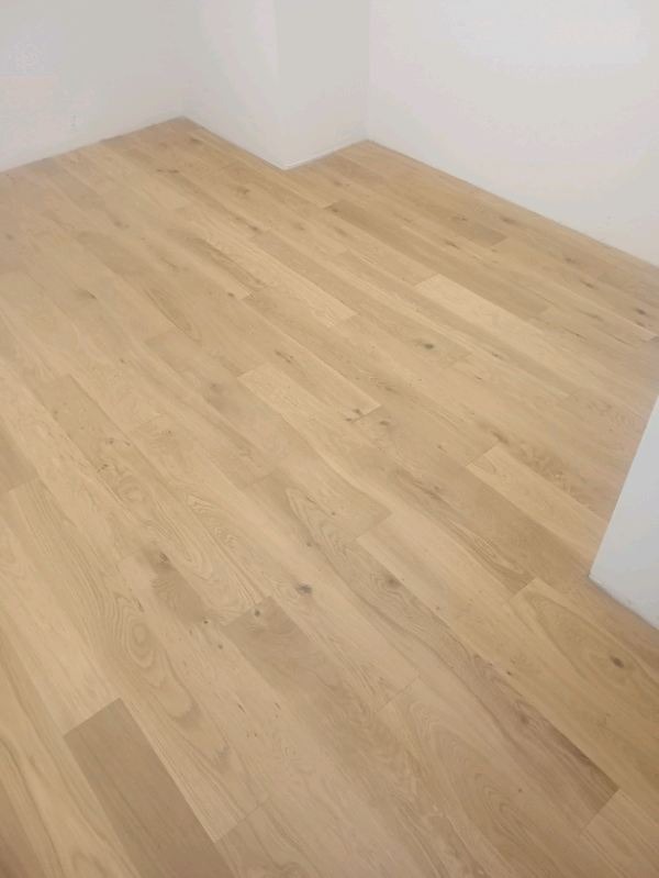 Jasna, drewniana podłoga ułożona w nowym, minimalistycznym wnętrzu. Widoczne subtelne usłojenie drewna i świeżo pomalowane, białe ściany.