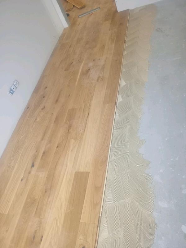 Układanie parkietu w jodełkę: widoczny fragment ułożonej podłogi z jasnego drewna i świeżo nałożony klej w charakterystyczny wzór. Praca w toku.