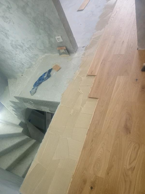 Układanie parkietu na schodach z użyciem kleju. Widoczne drewniane deski i spiralne schody w surowym stanie. Prace wykończeniowe w trakcie realizacji.