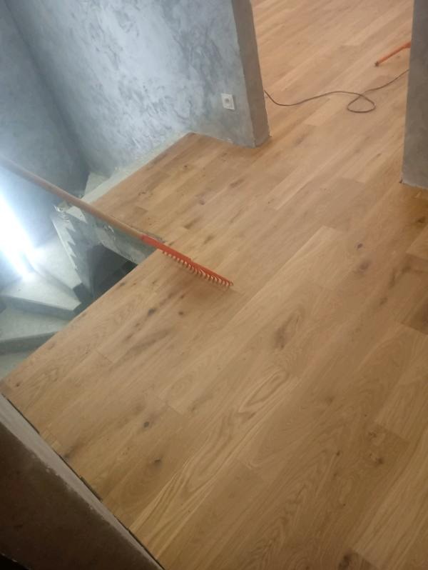 Ułożona drewniana podłoga w jasnym odcieniu, częściowo widoczne schody i szara ściana z gniazdkiem. W tle widoczne kolejne pomieszczenie.