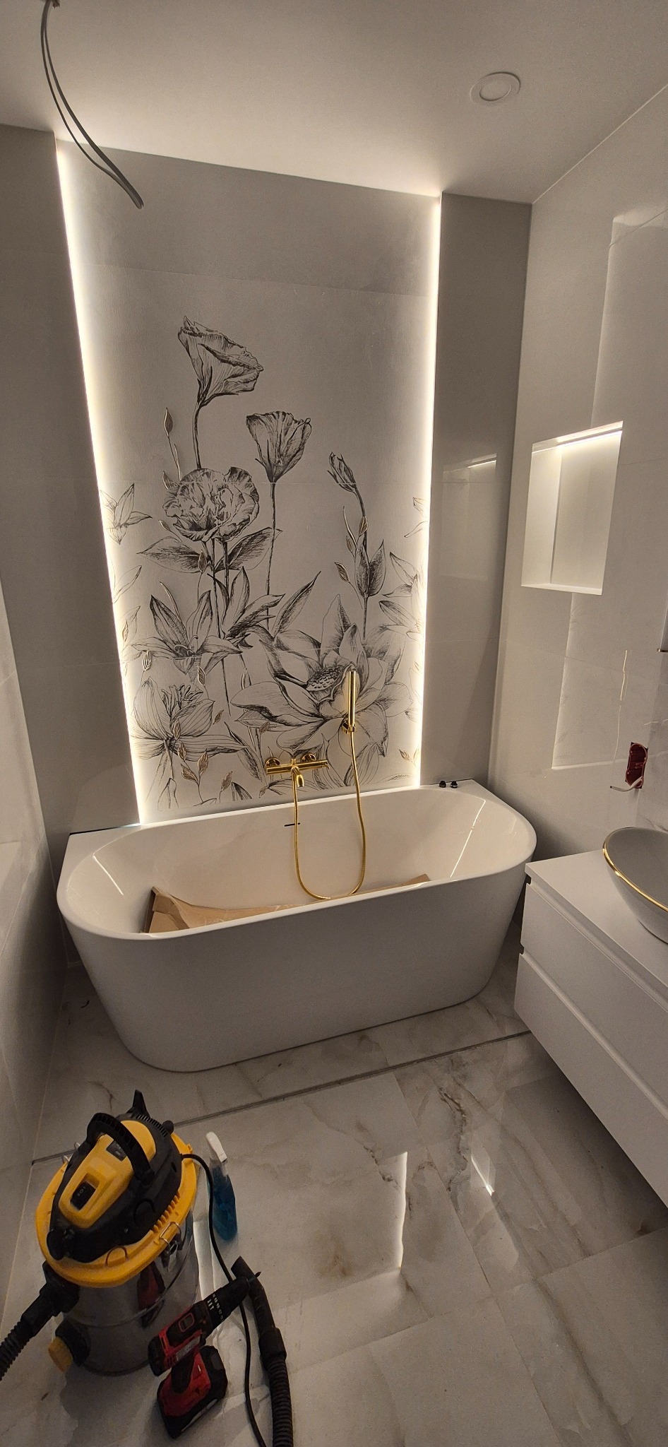 Nowoczesna łazienka z wolnostojącą wanną, złotą armaturą i podświetlaną ścianą z motywem kwiatowym. Na podłodze widoczny odkurzacz i narzędzia.