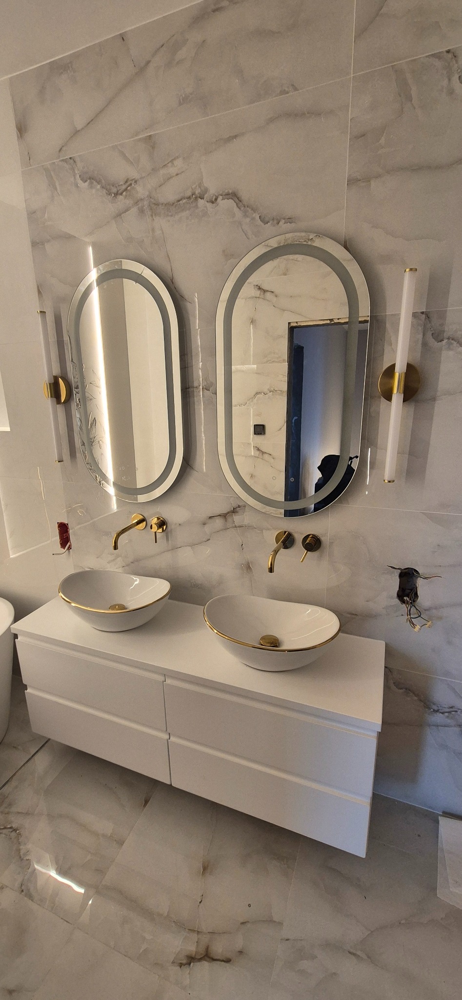 Wykończona łazienka z marmurowymi płytkami, owalnymi lustrami z podświetleniem LED, białymi umywalkami nablatowymi z złotymi akcentami i białą szafką pod umywalki.