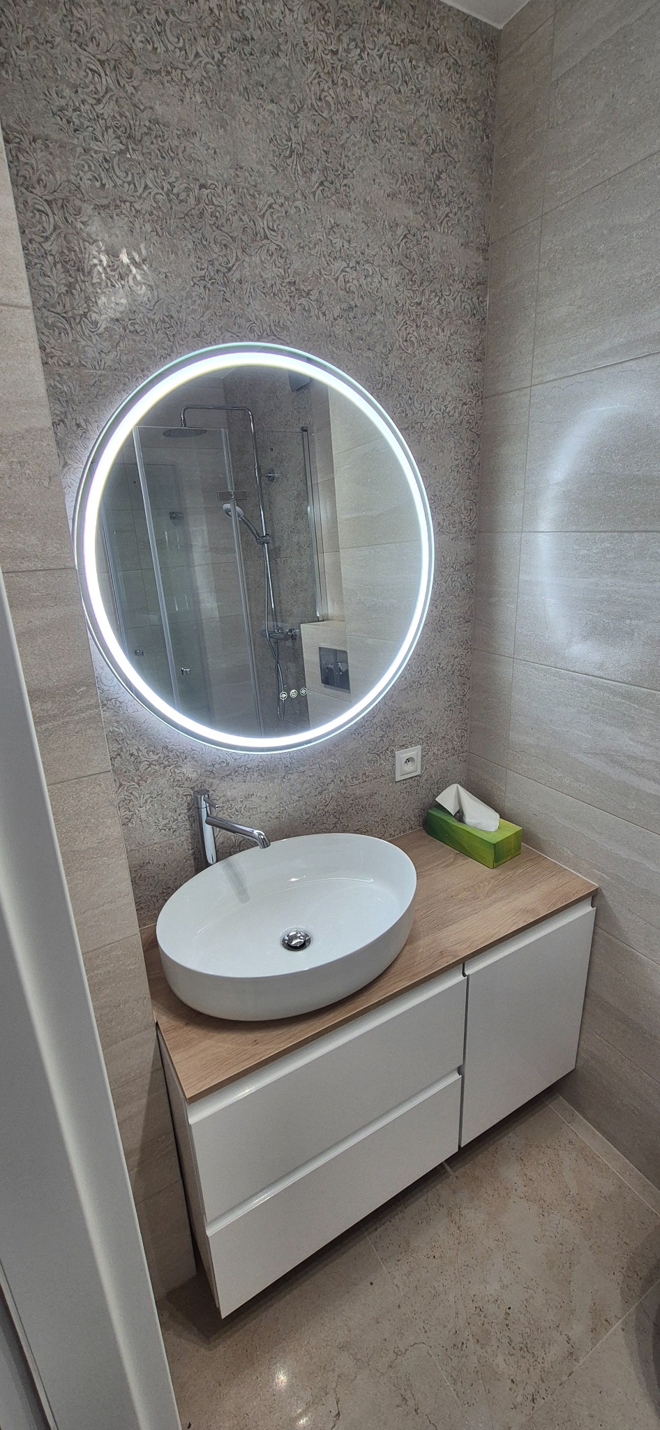 Wykończona łazienka w Łodzi: owalne umywalka nablatowa, szafka z drewnianym blatem i okrągłe lustro z podświetleniem LED. Eleganckie płytki na ścianach i podłodze.