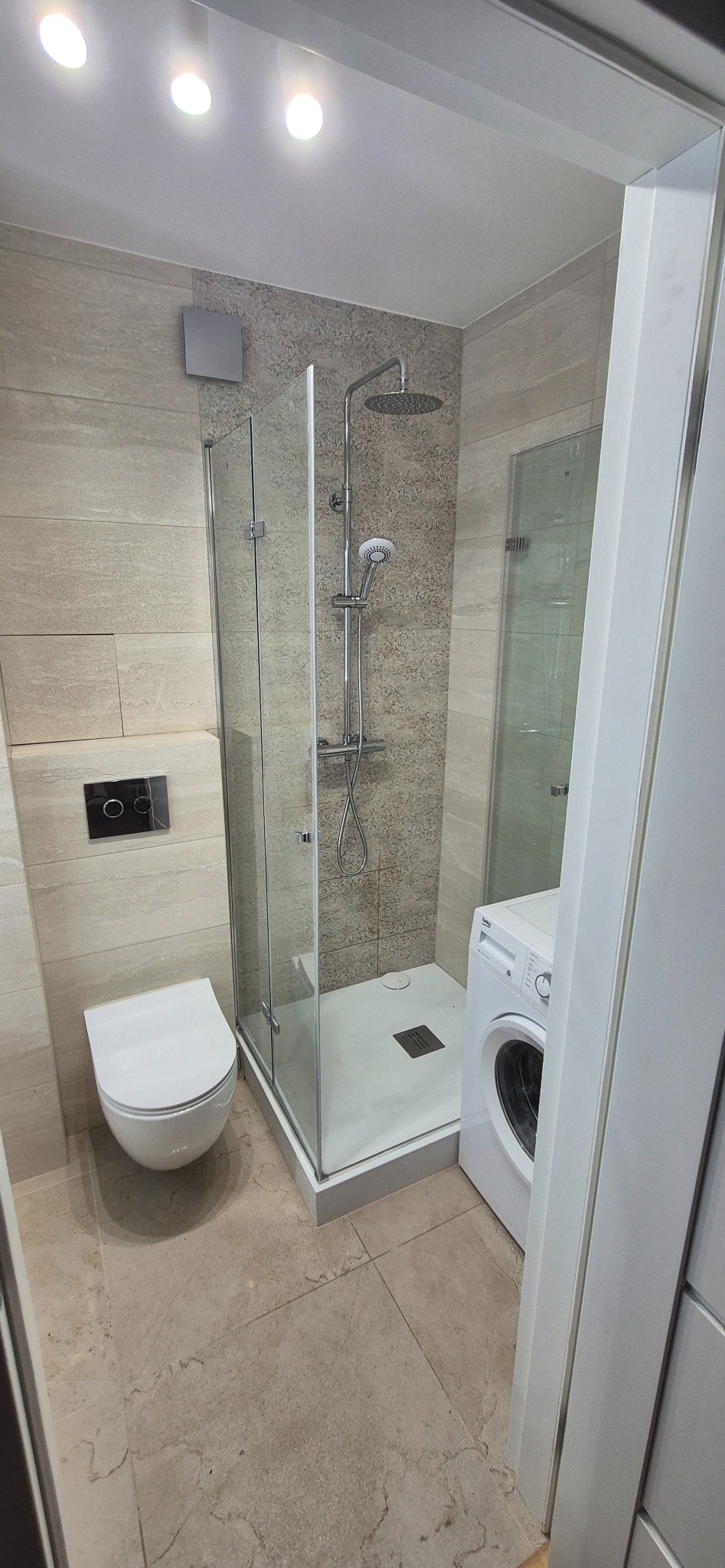 Jasna łazienka po remoncie: kabina prysznicowa ze szklanymi drzwiami, toaleta podwieszana, pralka. Płytki imitujące kamień na ścianach i podłodze. Oświetlenie punktowe.
