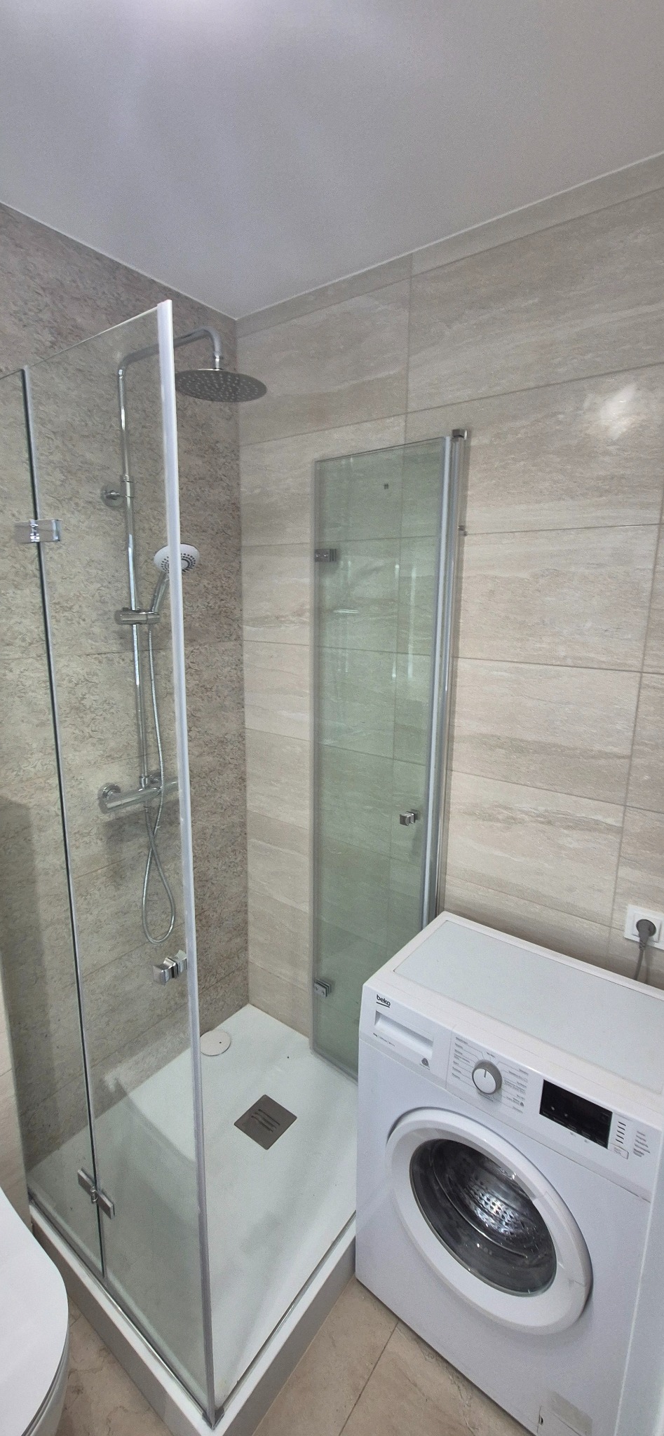 Wykończona łazienka z kabiną prysznicową typu walk-in, beżowe płytki imitujące kamień i biała pralka. Nowoczesny design, minimalistyczne wykończenie.