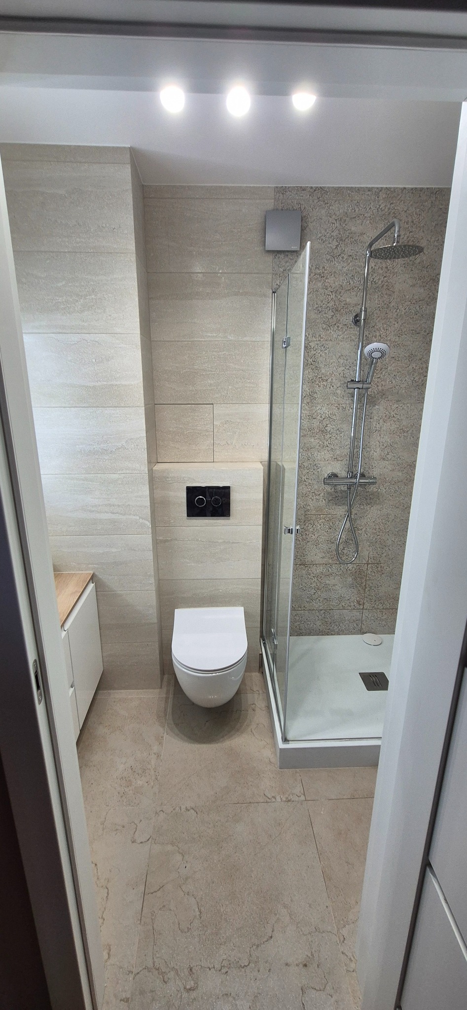 Jasna łazienka po remoncie: beżowe płytki imitujące kamień na ścianach i podłodze, kabina prysznicowa ze szklanymi drzwiami, podwieszana toaleta z czarnym przyciskiem.