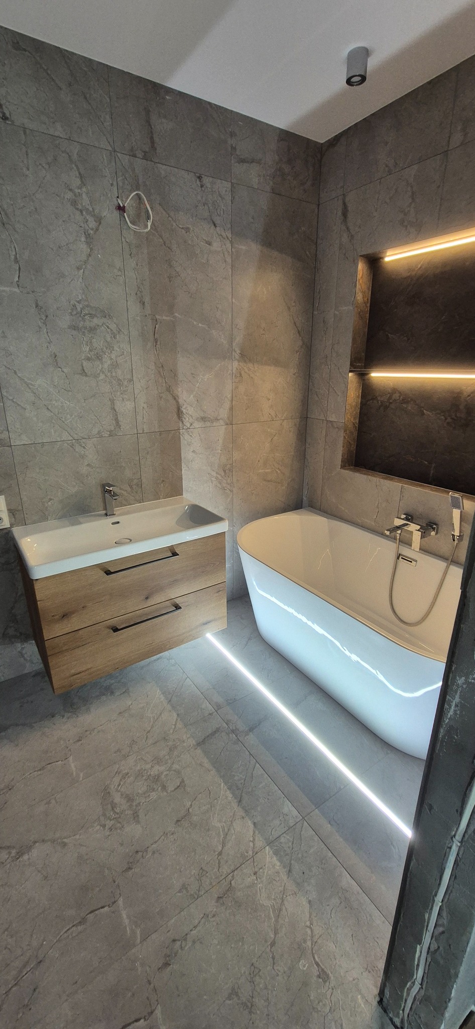 Nowoczesna łazienka z szarymi płytkami imitującymi marmur, drewnianą szafką pod umywalką i wolnostojącą wanną z podświetleniem LED. Minimalistyczny design.