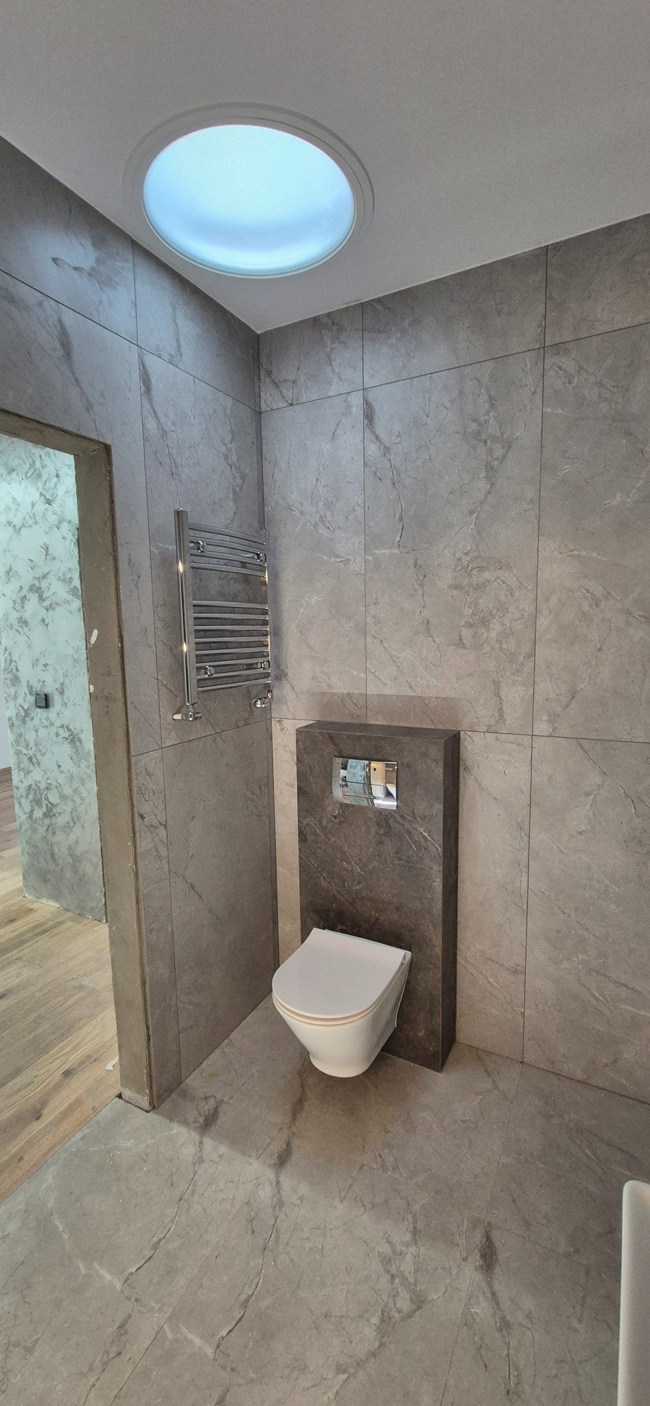 Nowoczesna łazienka z szarymi płytkami imitującymi marmur, podwieszana toaleta i okrągłe okno sufitowe. Grzejnik drabinkowy na ścianie.
