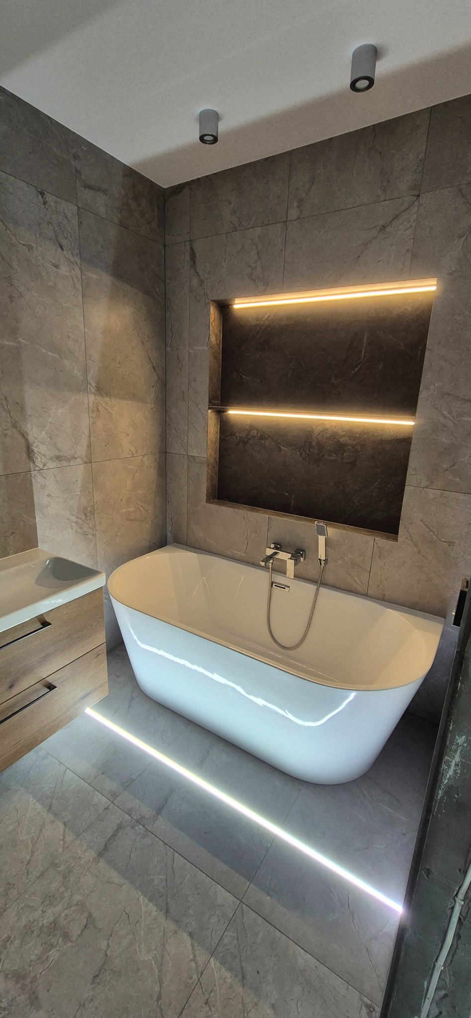 Nowoczesna łazienka z wanną wolnostojącą, szarą marmurową okładziną ścian i podświetlanymi wnękami. Minimalistyczny design, oświetlenie LED podkreśla elegancję.