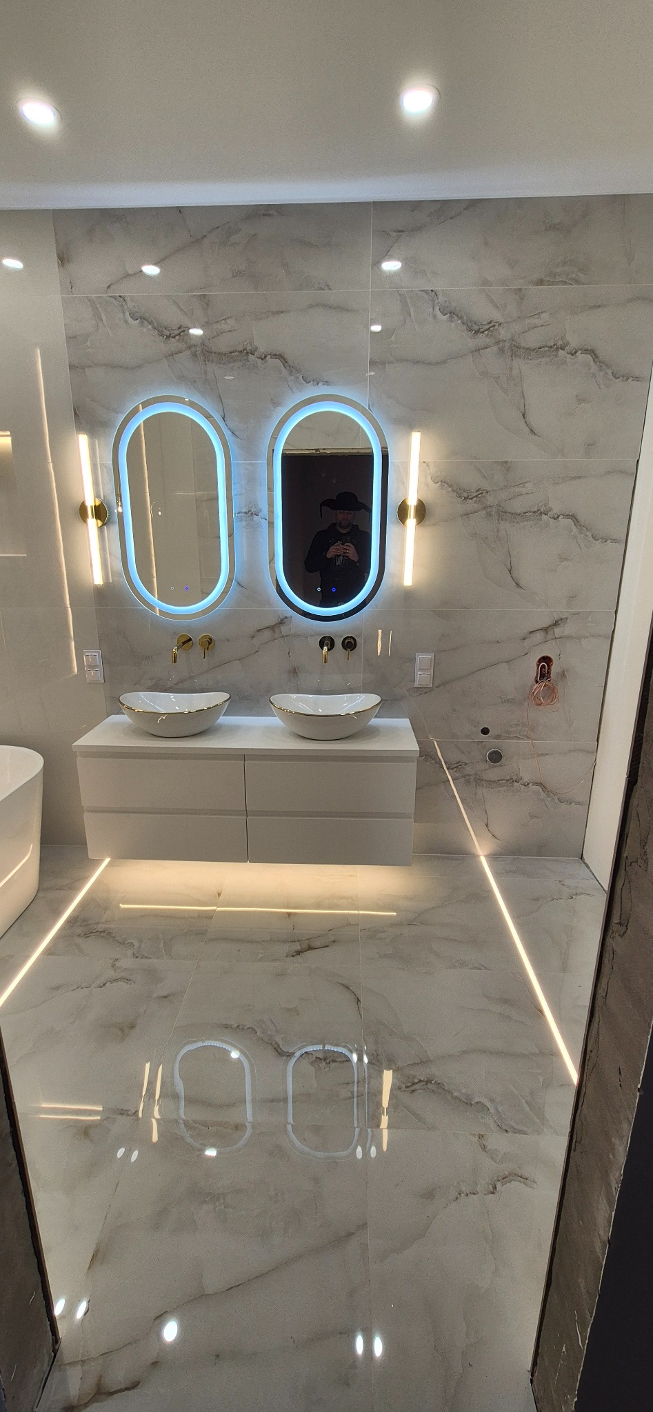 Nowoczesna łazienka z dwoma owalnymi lustrami LED, białymi umywalkami nablatowymi i marmurowymi płytkami. Widoczne złote detale armatury i oświetlenia.