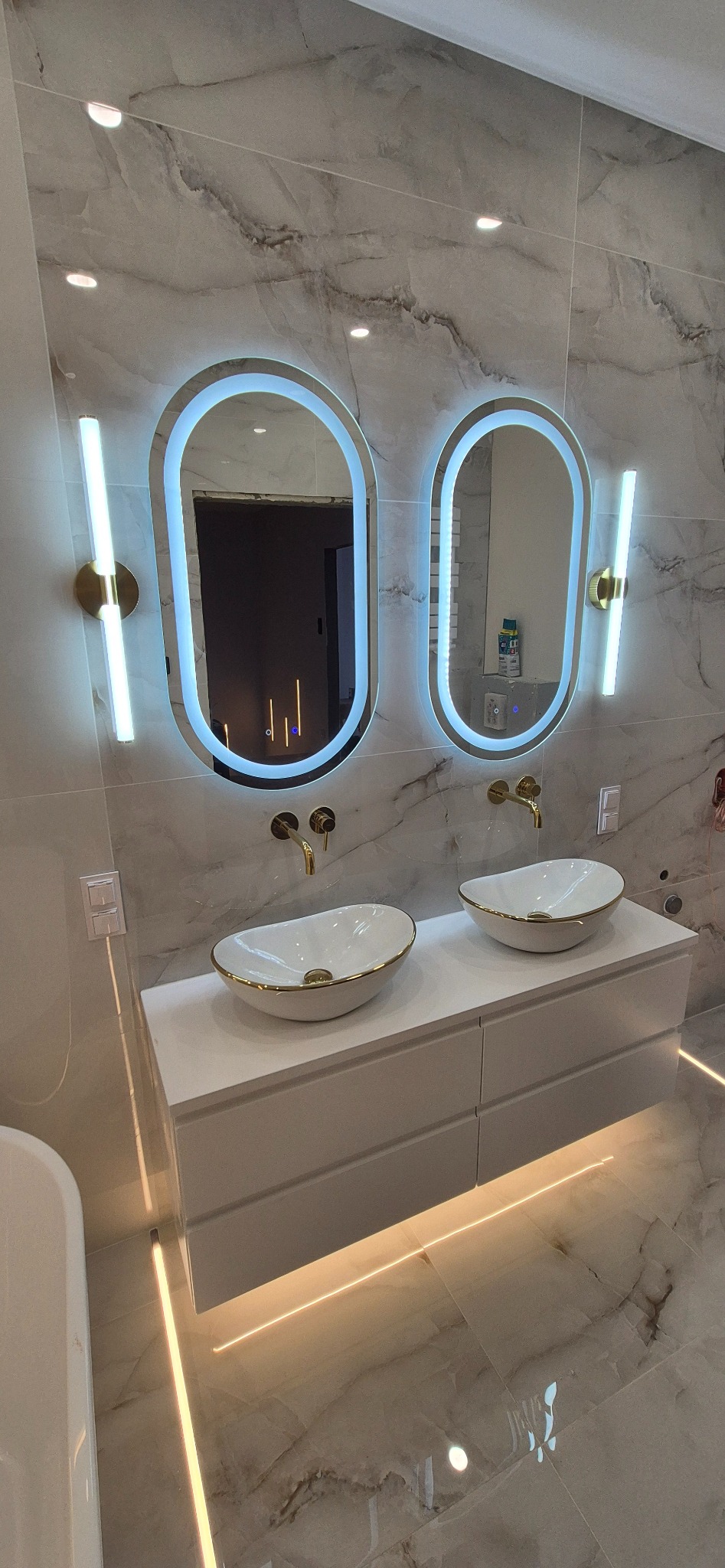 Nowoczesna łazienka z dwoma owalnymi lustrami LED, białymi umywalkami z złotymi akcentami i podświetlaną szafką na tle marmurowych płytek.