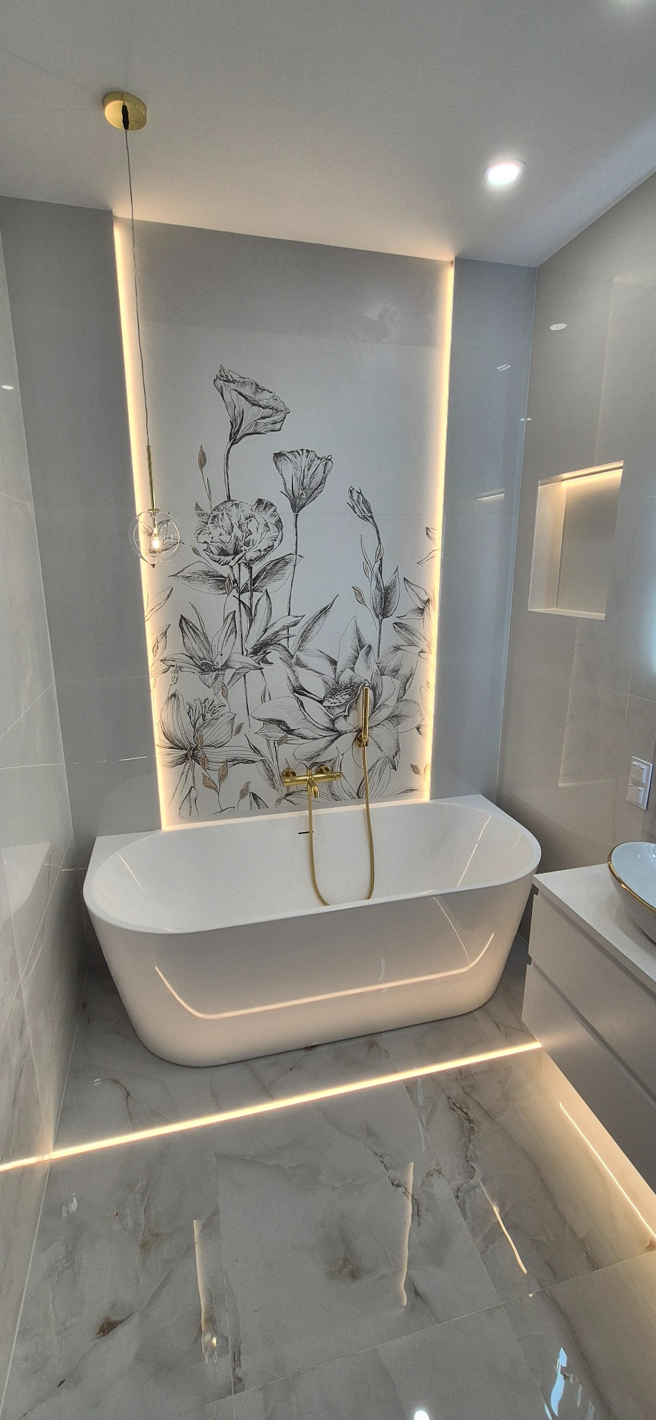 Nowoczesna łazienka z białą wanną, złotą armaturą i podświetlanym panelem z motywem kwiatowym. Marmurowe płytki podłogowe i subtelne oświetlenie LED.