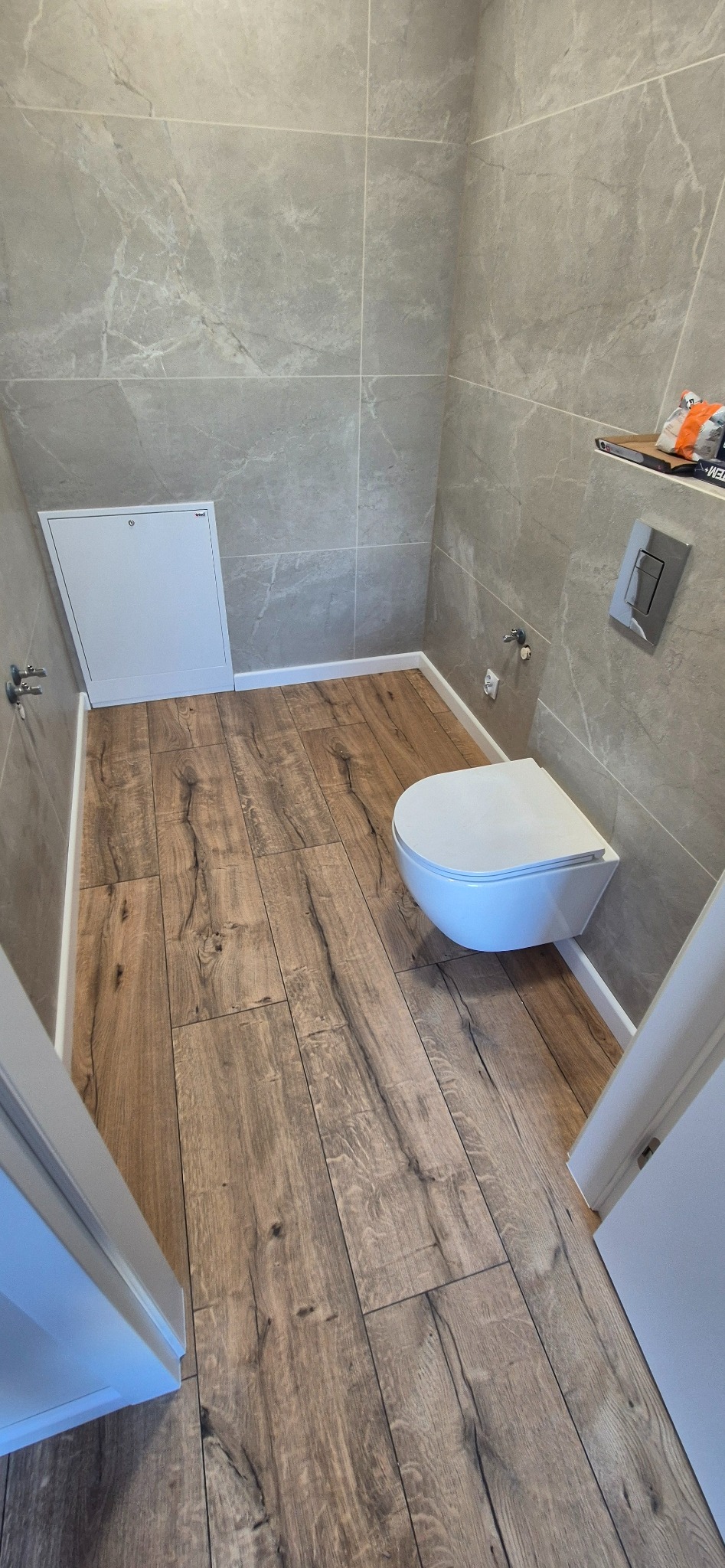 Nowoczesna toaleta z drewnopodobną podłogą i szarymi płytkami imitującymi marmur. Podwieszana miska WC i ukryty zawór. Minimalistyczny design.