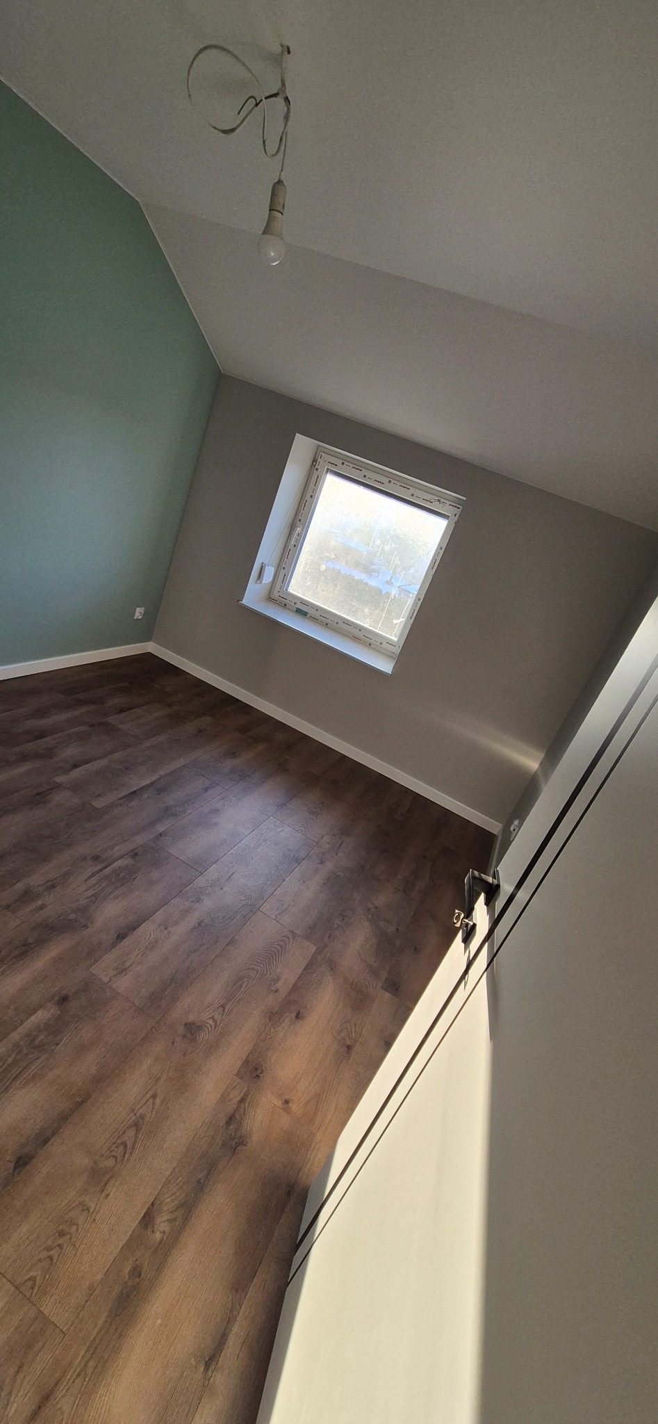 Pokój na poddaszu po remoncie: brązowe panele podłogowe, szare ściany, okno dachowe. Jedna ściana pomalowana na zielono. Widoczna żarówka na kablu.