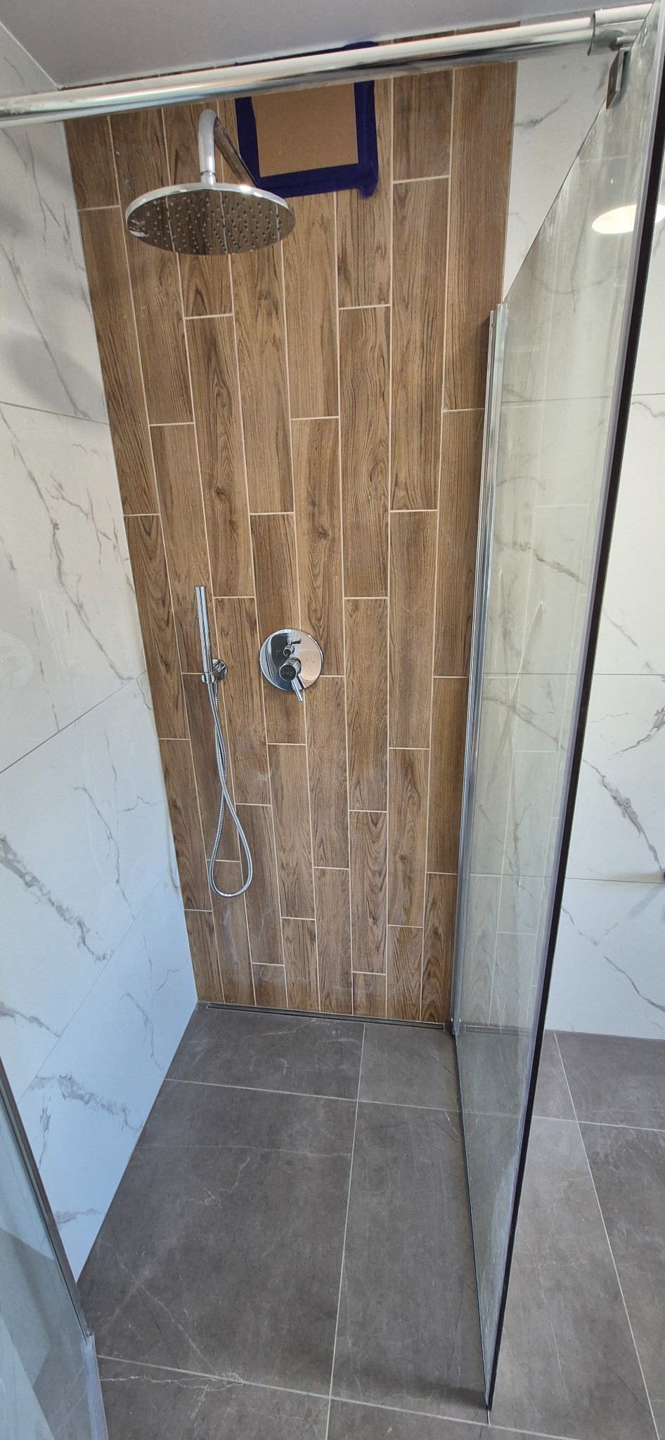 Wykończona kabina prysznicowa z drewnopodobnymi płytkami na ścianie, deszczownicą i szklanymi drzwiami. Szare płytki podłogowe z delikatnym wzorem marmuru.