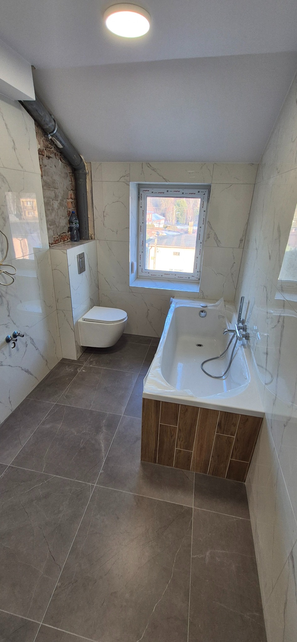 Wykończona łazienka z wanną w folii ochronnej, podwieszaną toaletą i szarą podłogą. Widoczne białe płytki imitujące marmur i fragment odsłoniętej cegły.